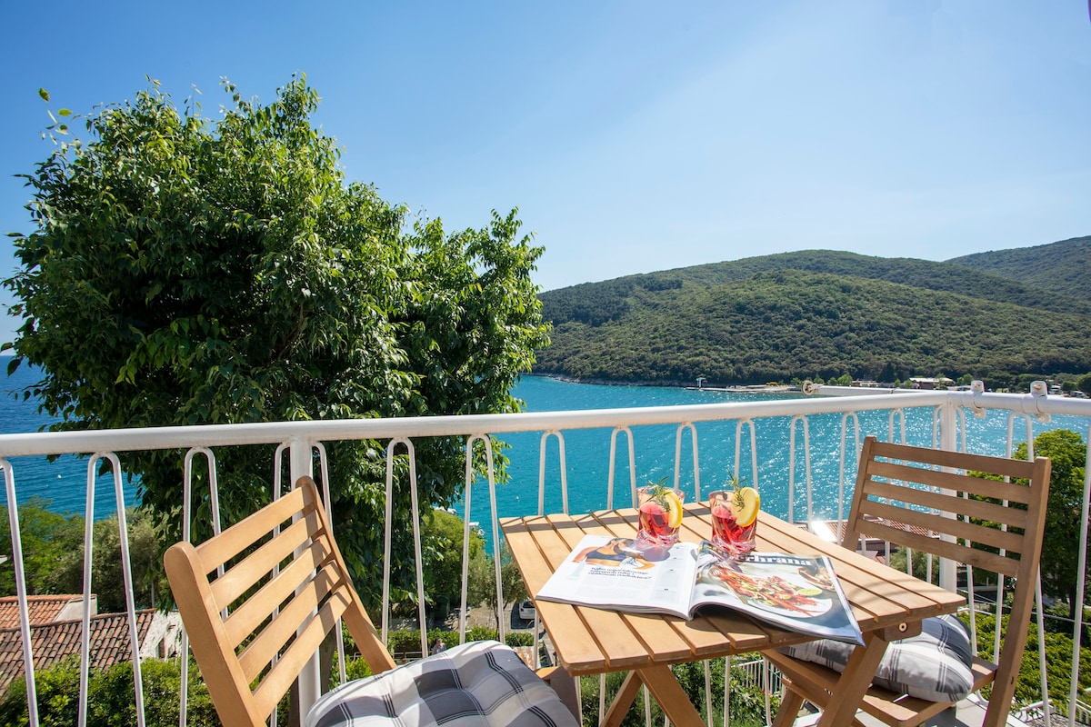 A balcony with a small wooden table and two matching chairs overlooks a scenic view of the sea and surrounding hills. Two drinks are placed on the table beside an open magazine, inviting relaxation in a peaceful outdoor setting.