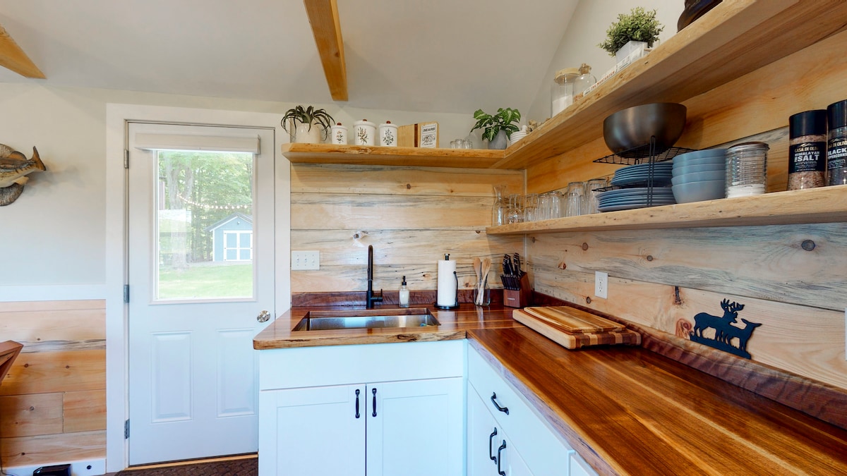 The kitchen features wooden shelving displaying glassware and dishes. A sleek sink area is complemented by a countertop made from rich, dark wood. Natural light from the door illuminates the space, highlighting the combination of wood textures in a warm, inviting environment.