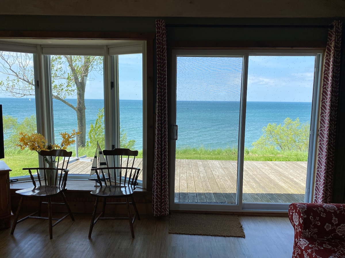 Large windows provide a panoramic view of the water, showcasing shades of blue. Two wooden chairs are positioned near the window, inviting relaxation. The deck outside extends towards the water, framed by trees that gently sway in the breeze.