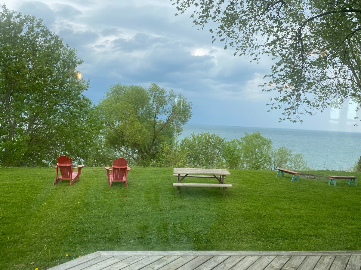 A spacious lawn borders the waterfront, featuring two red Adirondack chairs and a picnic table. Lush greenery surrounds the area, with a view of the serene lake beyond. The sky shows hints of clouds, indicating a peaceful atmosphere.