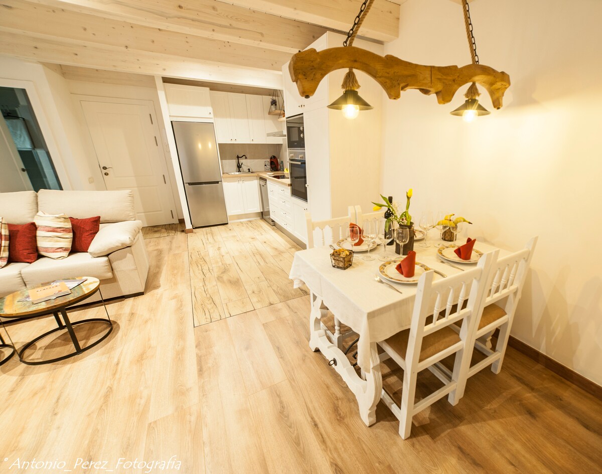 A cozy dining area features a white wooden table set for four, accompanied by matching chairs. A modern kitchen with stainless steel appliances is visible in the background, while a comfortable seating area is highlighted by a light-colored sofa and decorative pillows.