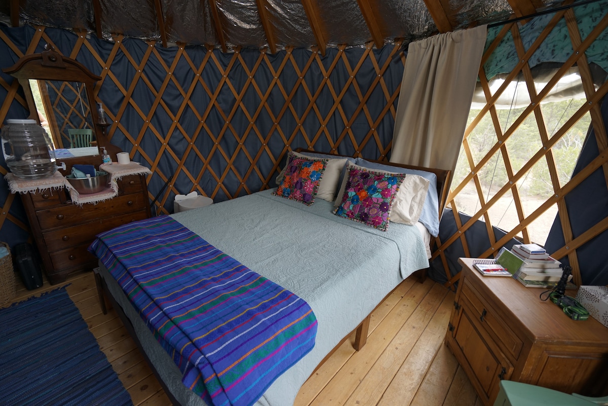 A spacious sleeping area features a neatly made bed draped in a light-blue coverlet. Colorful decorative pillows add a touch of vibrancy. A wooden dresser and bedside table complement the natural wooden flooring. Soft light filters through the window, accented by beige curtains.