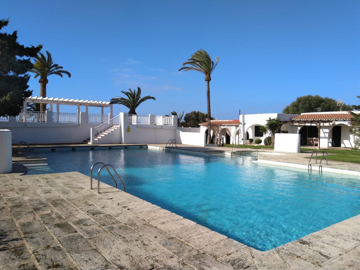 A community pool is showcased, featuring clear blue water and gently sloping steps for access. Surrounding palm trees and landscaped greenery add to the environment. A white terrace with shaded seating overlooks the pool area, creating a tranquil outdoor setting.
