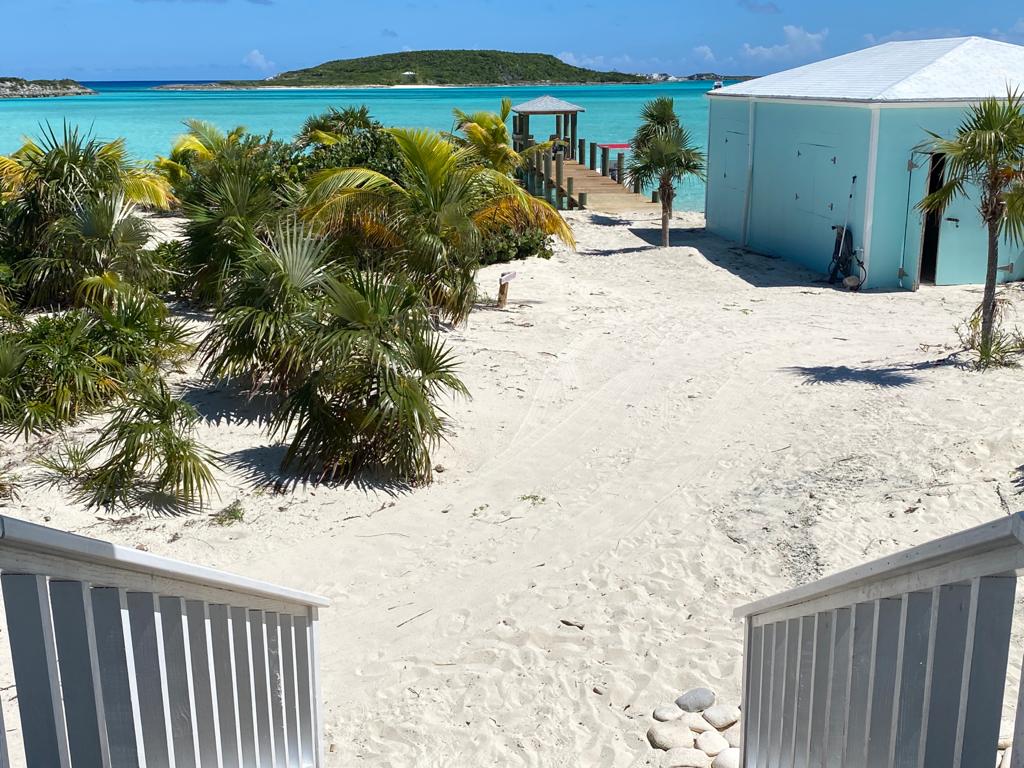 A view of sandy ground leading to a serene beachfront, featuring lush palm trees and turquoise waters in the background. The scene captures tropical natural beauty, with a dock visible to the left and a light-colored building on the right.