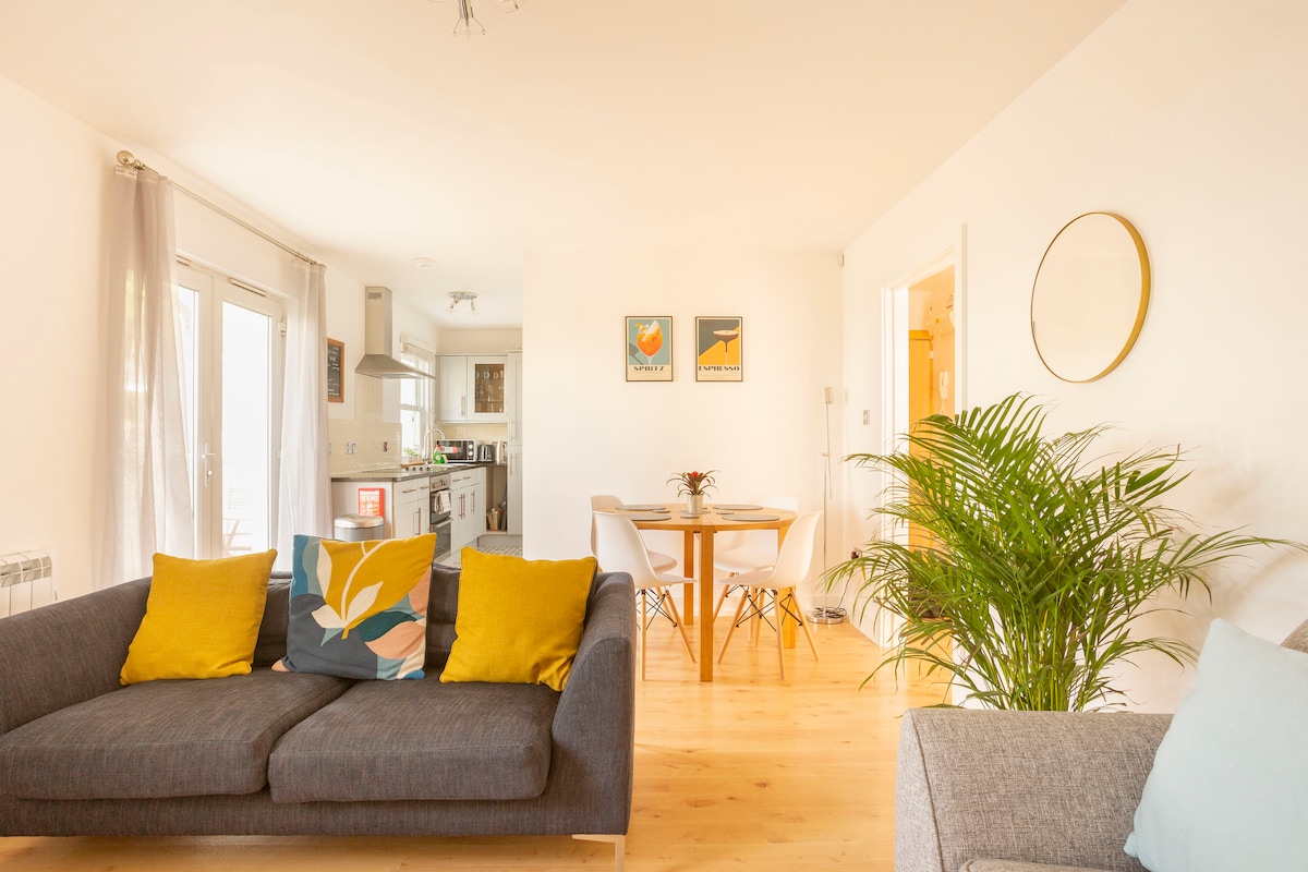 A spacious living area features two comfortable sofas accented with colorful cushions. A dining table for four is positioned nearby, illuminated by natural light. The kitchen area is visible in the background, complemented by minimalist decor and a vibrant indoor plant.