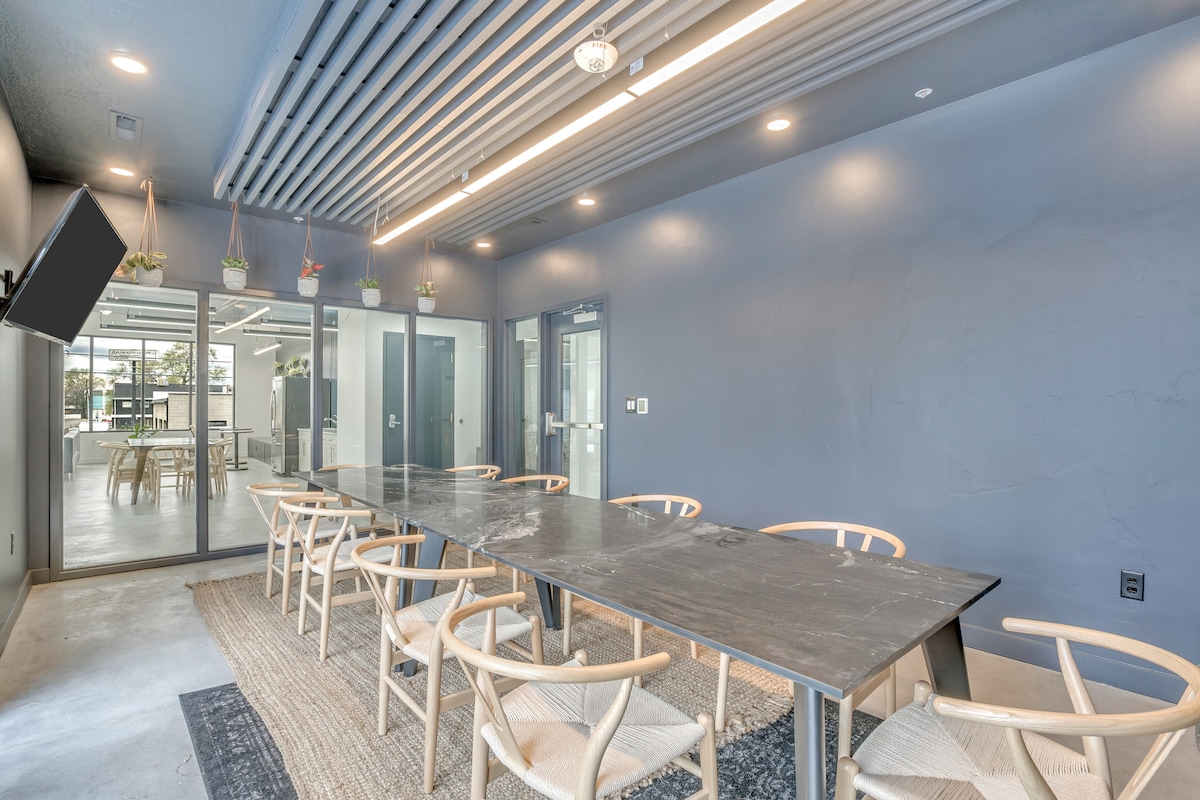 A coworking space is depicted with a long, rectangular table surrounded by multiple light-colored chairs. The walls are painted in a calming shade of blue, and large windows allow natural light to flood the area. Plants are displayed above the windows, adding a touch of greenery.