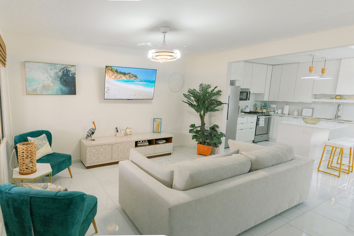A modern living area features a spacious light-colored sofa and two teal accent chairs, providing an open and inviting space. A flat-screen television is mounted on the wall above a minimalistic media console. A potted plant adds a touch of greenery near the kitchen entrance.
