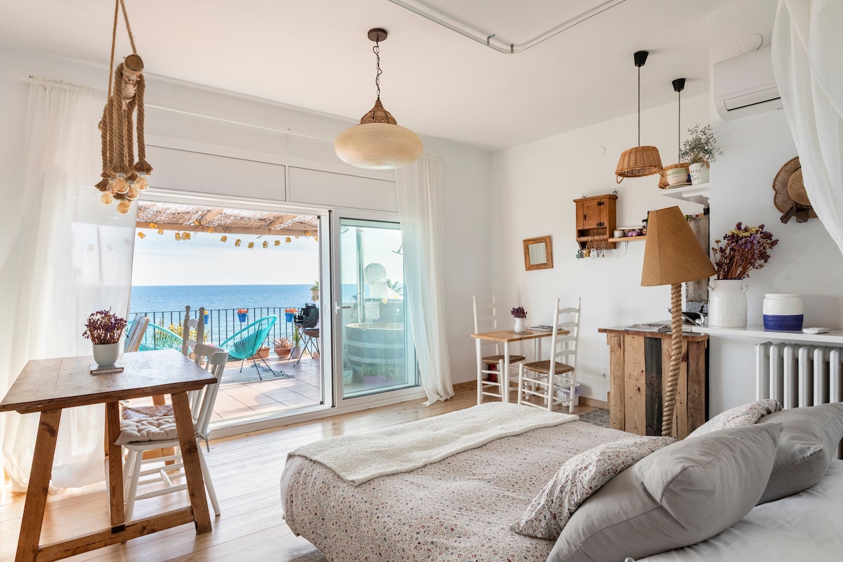 A bright and airy space features a spacious bed with soft linens, complemented by a dining table and chairs near a large glass door. The balcony presents a view of the sea, with decorative elements enhancing the room's coastal ambiance.