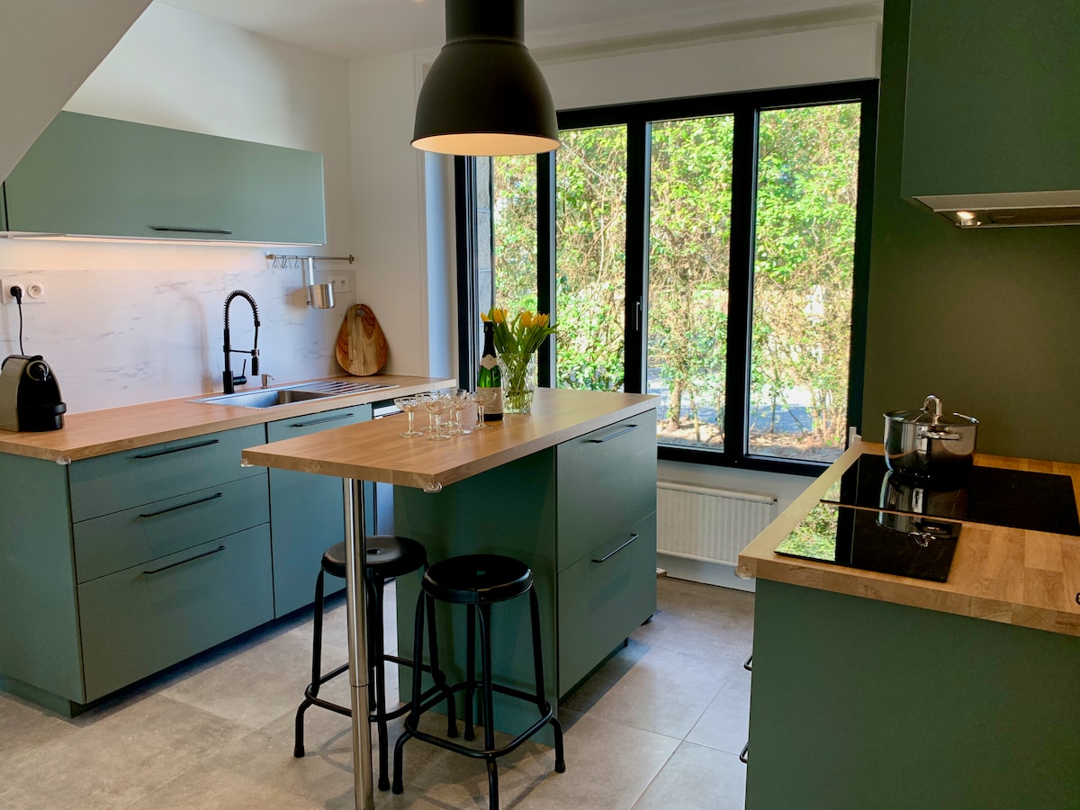The modern kitchen, featuring a green color scheme, includes sleek cabinetry and a central island with bar stools. Large windows provide natural light and a view of the outdoor greenery. An induction stove and countertop appliances are also visible, enhancing functionality.