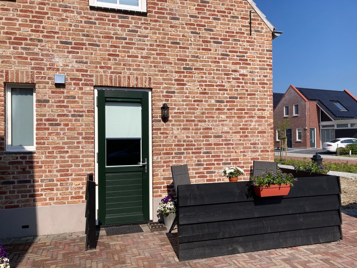 An exterior view showcases a brick facade of the studio, featuring a green door and a welcoming seating area with two chairs. A planter with blooming flowers adds a touch of color, while nearby properties are visible in the background under a clear blue sky.
