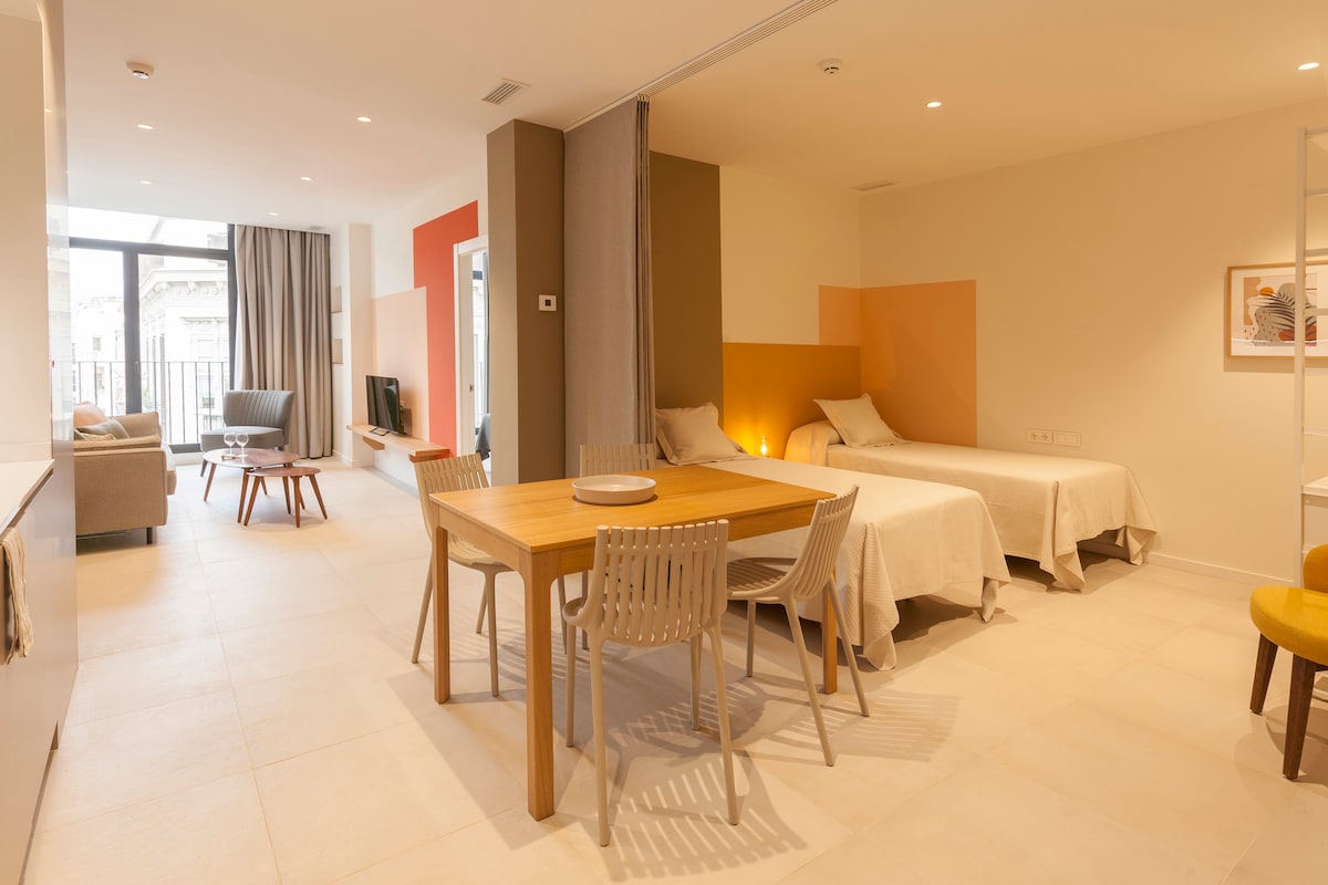 A modern studio apartment features a comfortable seating area with a television alongside a dining table surrounded by four chairs. Two spacious beds are neatly arranged, with soft lighting accentuating the room’s warm tones. Large windows allow natural light to fill the space.