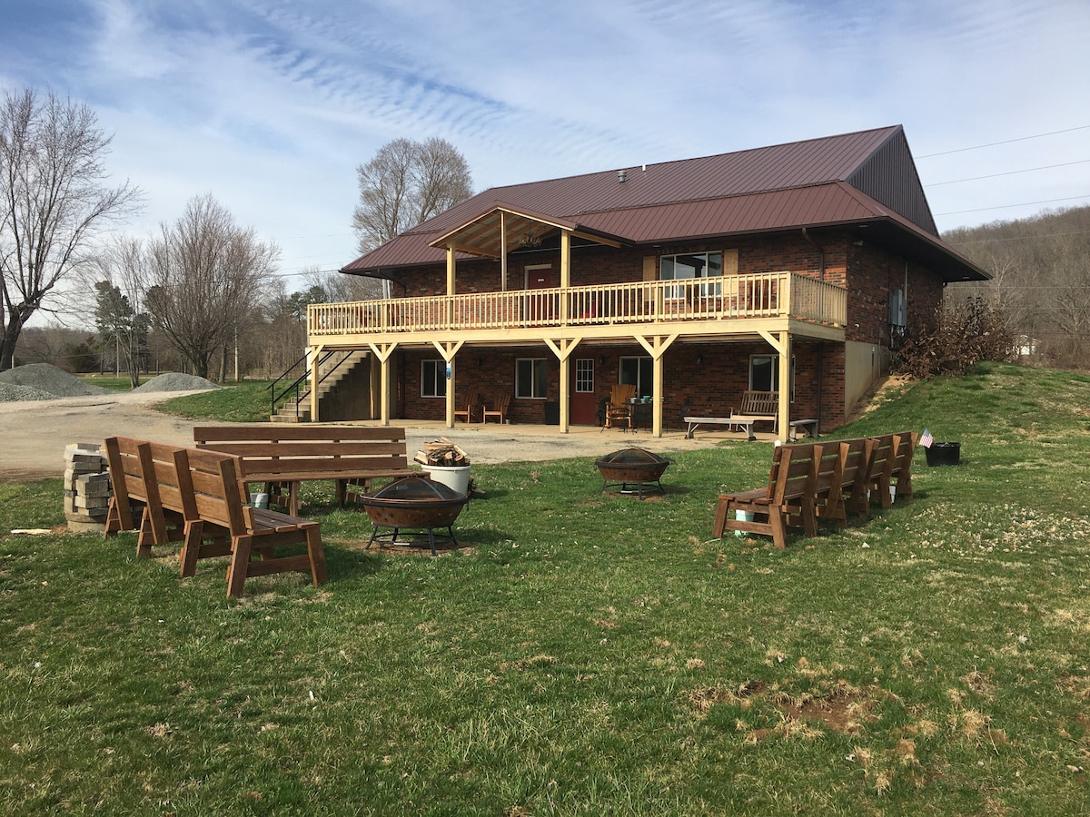 An outdoor area features several wooden benches arranged around multiple fire pits, situated on a grassy lawn. A rustic lodge, with a covered balcony, stands in the background. The surrounding landscape includes trees and a gentle slope.