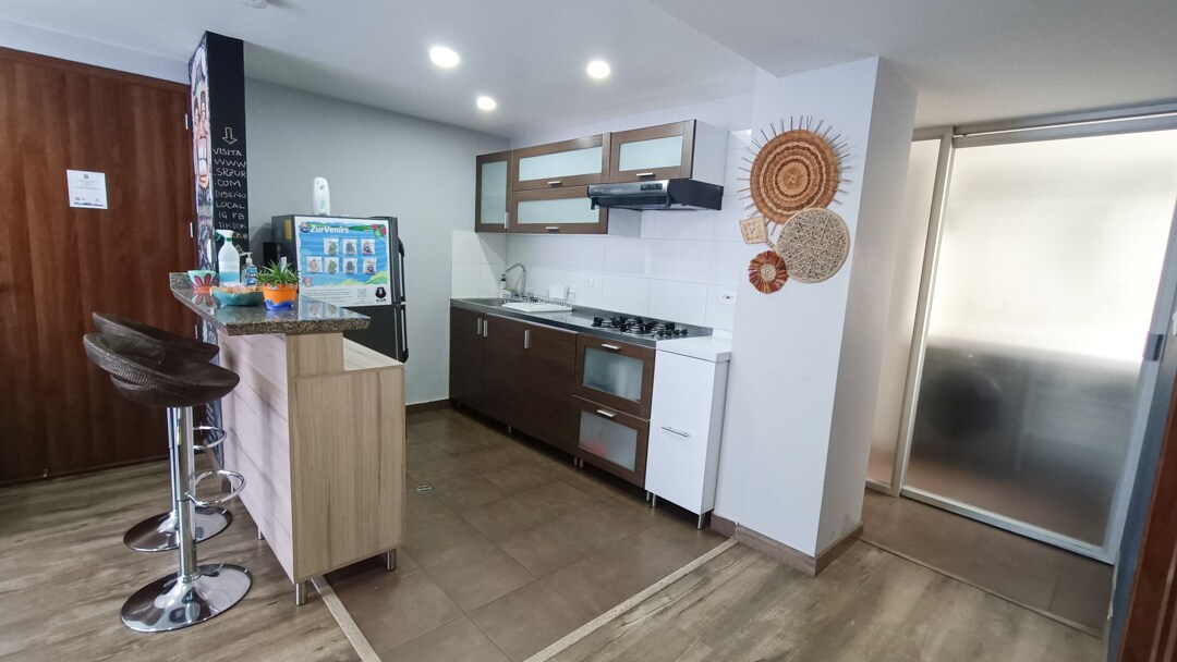 A modern kitchen area features sleek cabinetry and a gas stove. Bar stools are positioned at a counter, while a refrigerator stands nearby. Decorative elements, including round wall art, add character. The space is well-lit with overhead lighting and an open layout leading to the adjacent room.