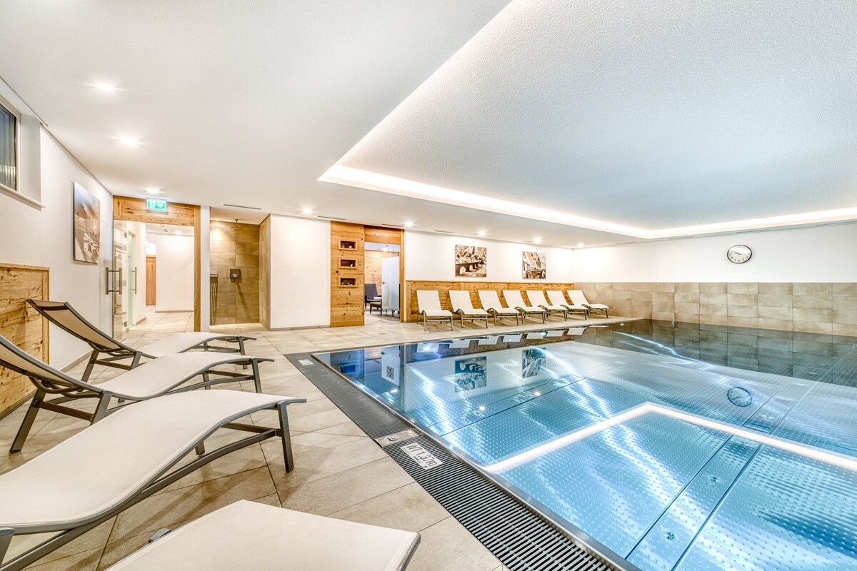 An indoor pool area features a tranquil swimming pool surrounded by modern lounge chairs. Soft ambient lighting highlights the space, which includes a clock on the wall and natural wood accents. The clean lines and muted color palette contribute to a relaxing atmosphere.