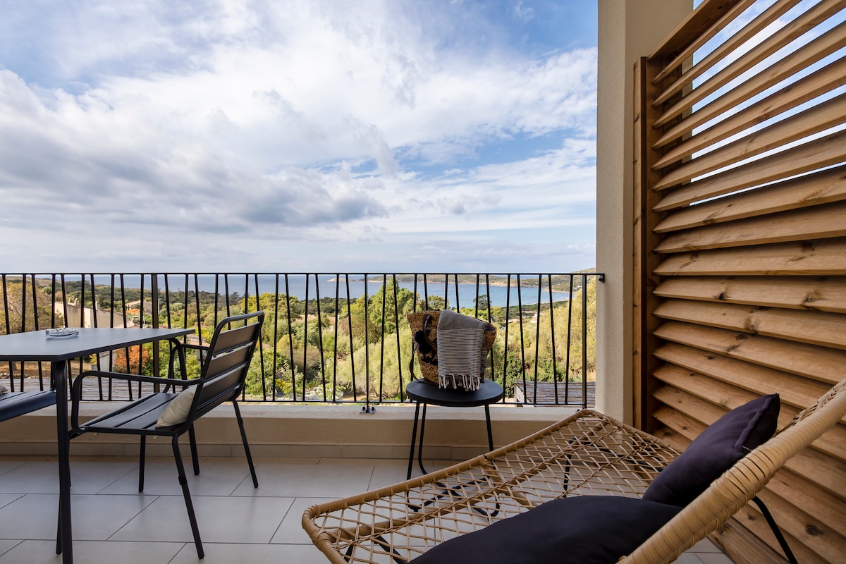 A terrace with a view overlooking lush greenery and the coastline. A dining table and chairs are positioned alongside a lounge chair. The balcony rail features wooden slats, providing a sense of privacy while allowing light to filter through.
