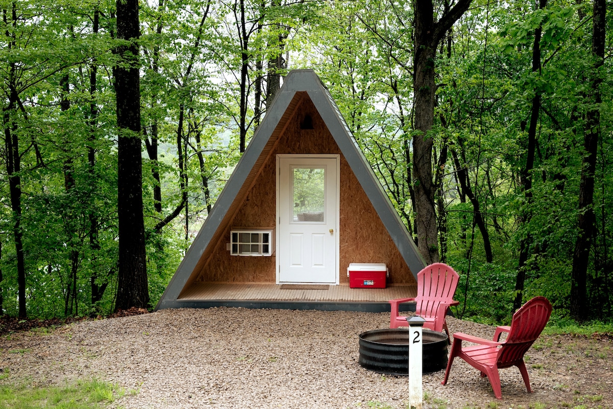 An A-frame cabin is nestled among lush green trees, featuring a clean white door and a small air conditioning unit beside it. Two red Adirondack chairs are positioned around a circular fire pit on gravel, offering a cozy outdoor gathering space.