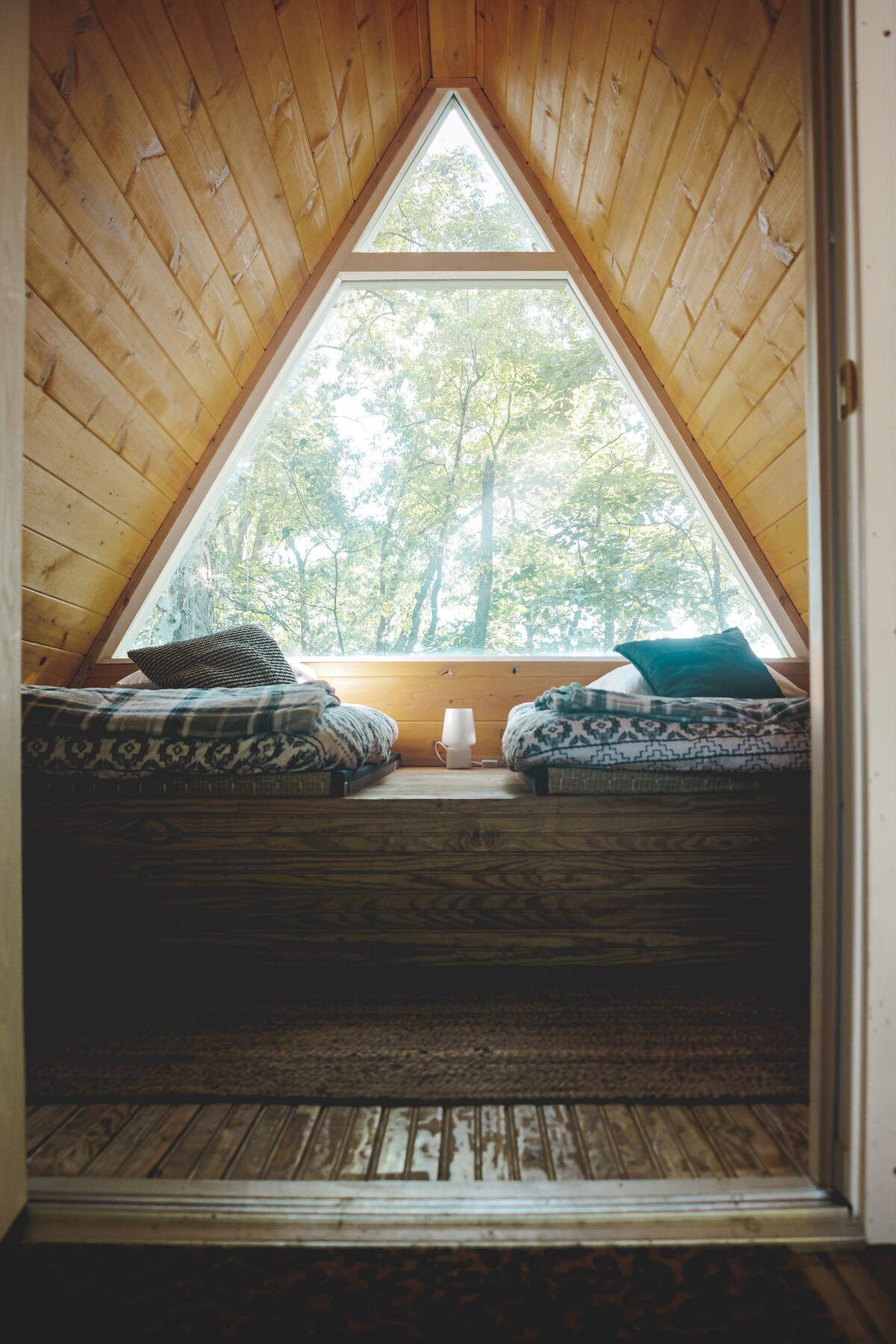 A serene corner of the A-Frame cabin features two comfortable seating areas by a large triangular window. Natural light fills the space, illuminating the wooden walls and creating a peaceful atmosphere. Decorative cushions add a touch of coziness to the inviting nook.