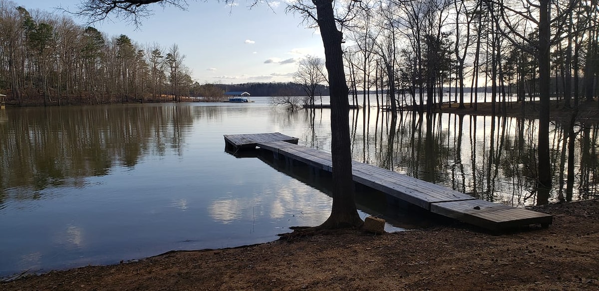 A wooden dock extends into calm waters, surrounded by trees reflecting on the surface. The landscape captures the serene atmosphere of Kerr Lake, with a distant boat visible in the background, framed by a clear sky and gentle clouds.