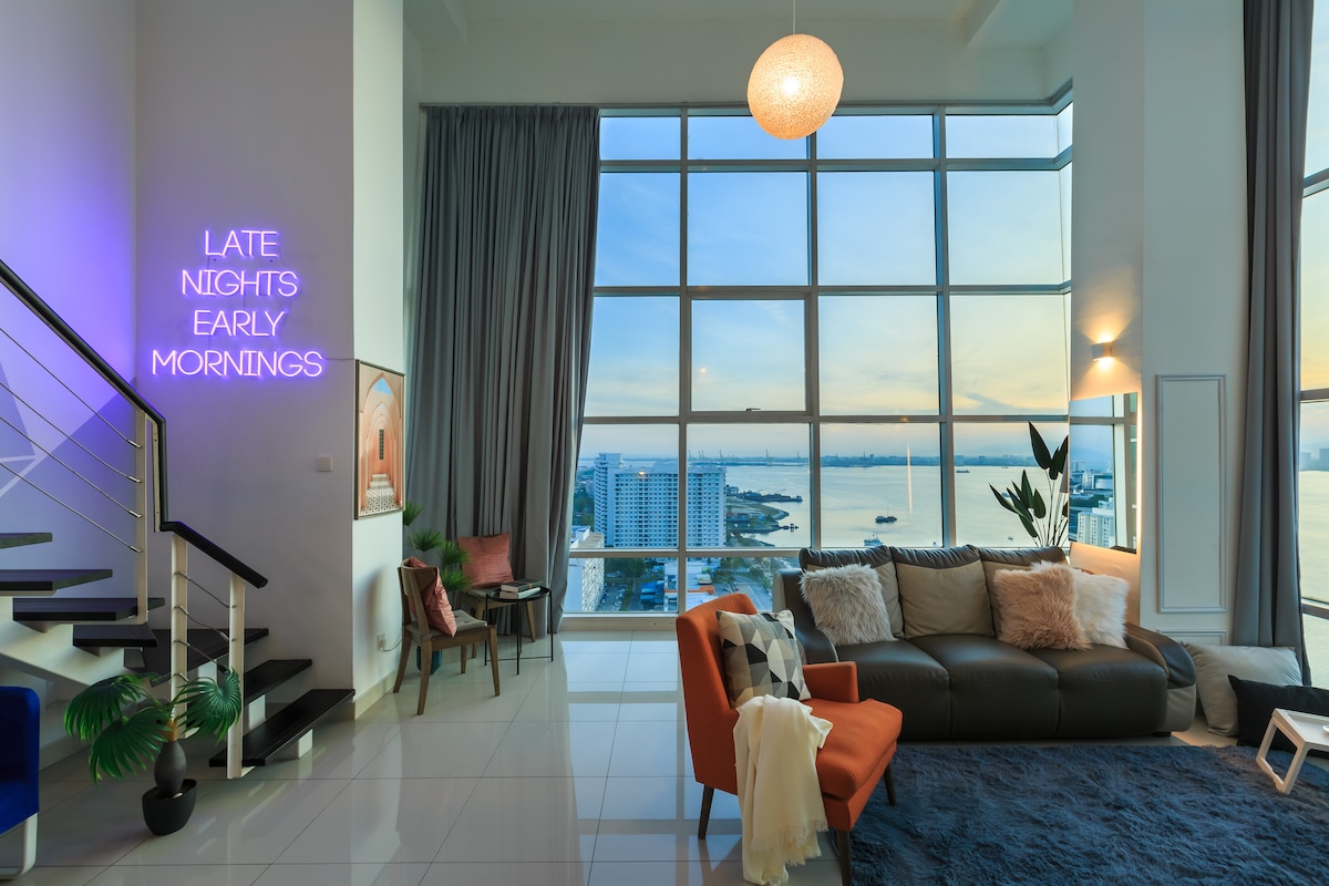 A spacious living area is highlighted by large windows offering a stunning view of the seafront. A modern sofa, adorned with soft pillows, is complemented by an accent chair. A statement light fixture hangs above, and a neon sign on the wall adds a unique touch.