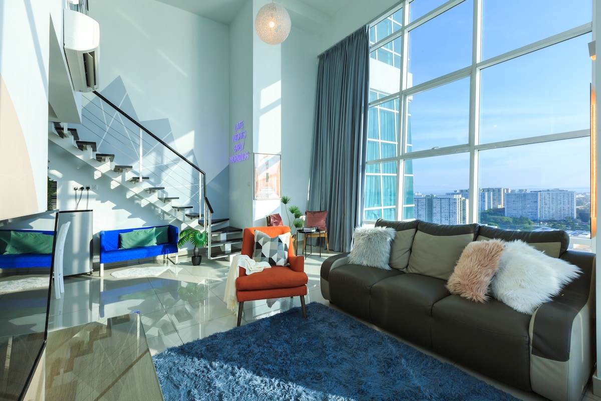 A spacious living area features large windows that allow natural light to flood the space. A comfortable sofa with soft throws faces a plush area rug. The room is accented with modern decor, including a vibrant armchair and contemporary plants. A staircase is visible in the background.