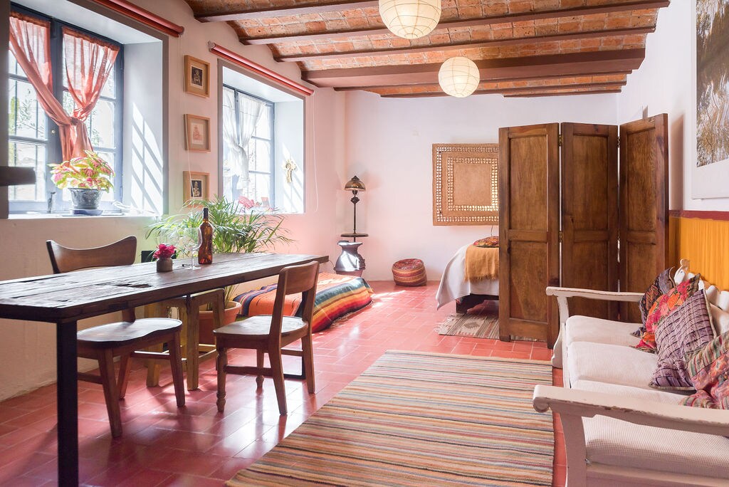 A welcoming living space is characterized by wooden beams and tiled flooring. A dining table with wooden chairs is positioned adjacent to a cozy seating area featuring colorful textiles. Natural light streams through large windows, illuminating decorative plants and art adorning the walls.