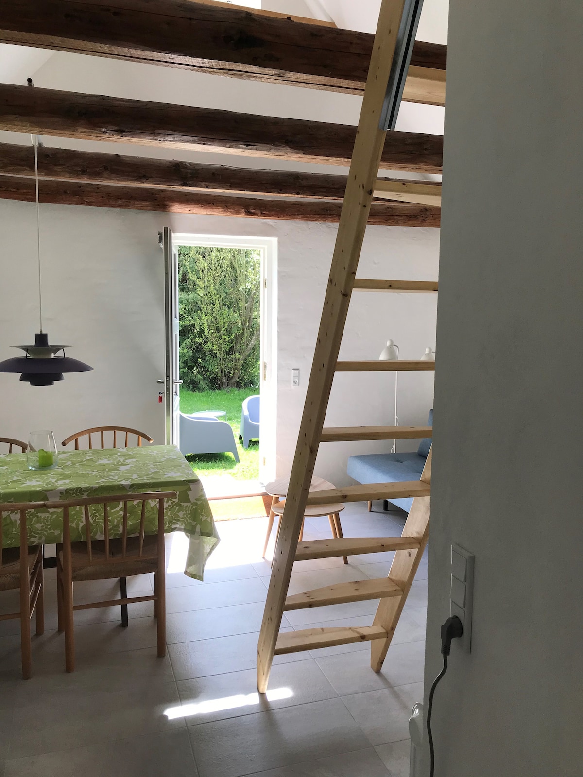 A wooden ladder leads to a cozy loft, while a dining area is configured with a light-colored table and four chairs. A glass door opens to a green outdoor space, complemented by a comfortable sofa and a modern pendant light above the table.