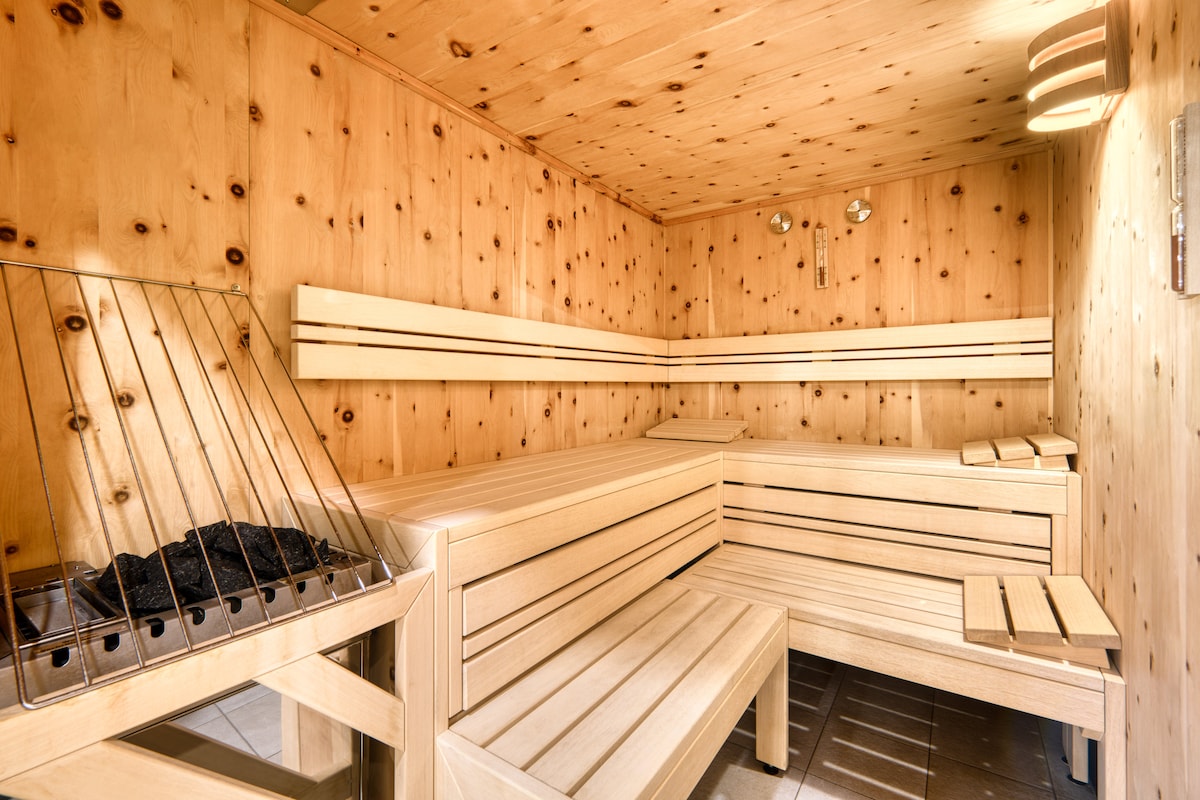 A wooden sauna features bench seating on multiple levels and a traditional sauna stove. The walls made of light-colored wood create a warm and natural ambiance, while soft lighting adds to the calming atmosphere.