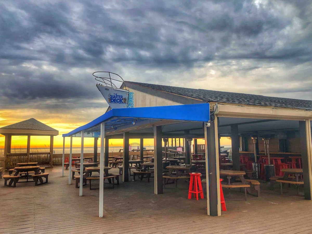 An open beachfront dining area features wooden picnic tables under a blue canopy. A wooden deck extends along the space, leading toward ocean views. The sky exhibits a mix of grey and orange hues at sunset, creating a calm atmosphere.