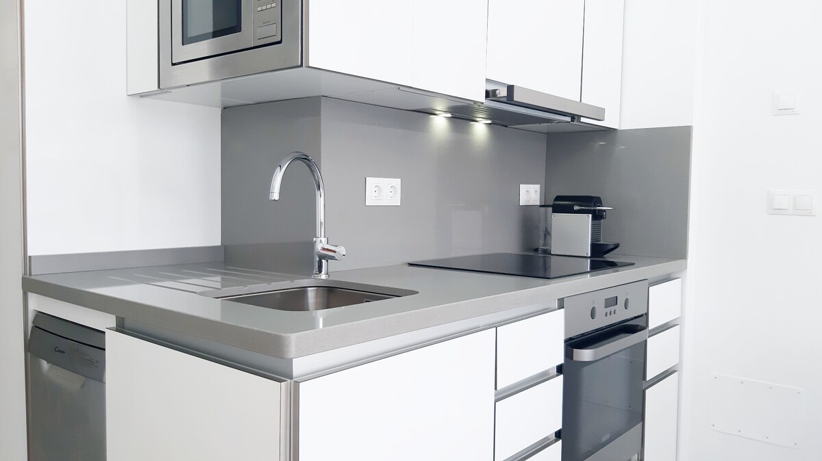 A modern kitchen is depicted with sleek white cabinetry and a light gray countertop. Stainless steel appliances include a built-in microwave, an oven, and a dishwasher. The kitchen features a single basin sink with a contemporary faucet, providing a clean and functional cooking space.