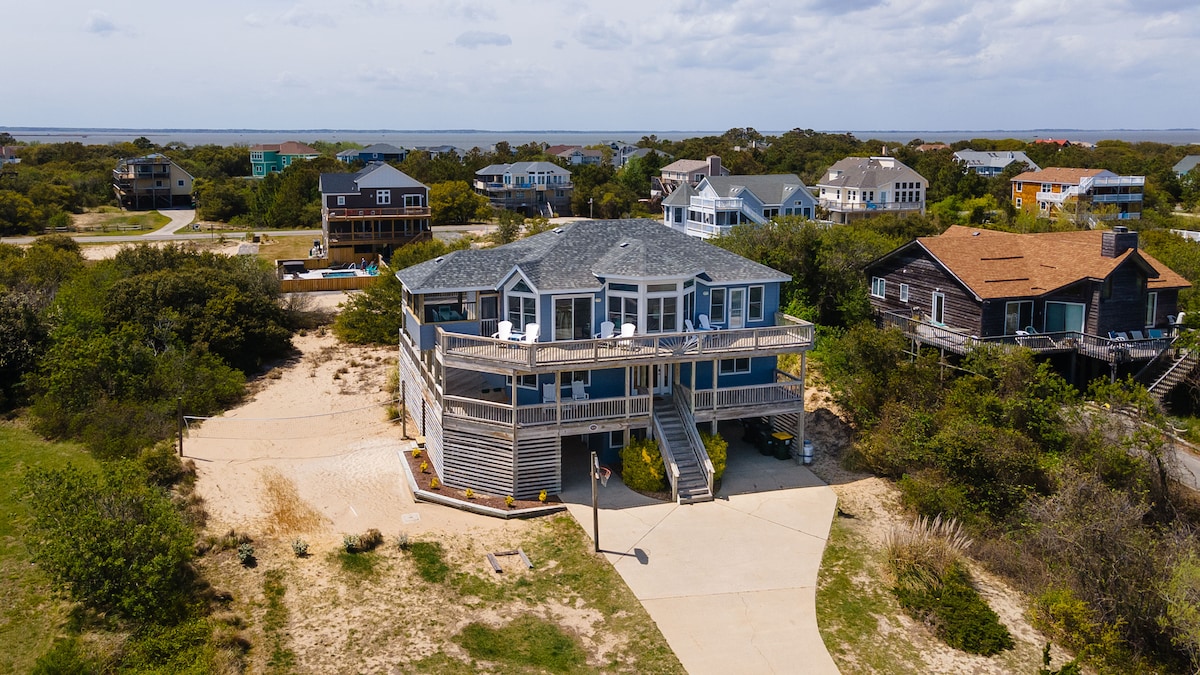The spacious beach house is nestled among lush greenery, with multiple levels of deck space visible. An expansive front porch welcomes guests, while ocean views can be seen in the distance. The surrounding area features a mix of residential homes and natural landscapes.