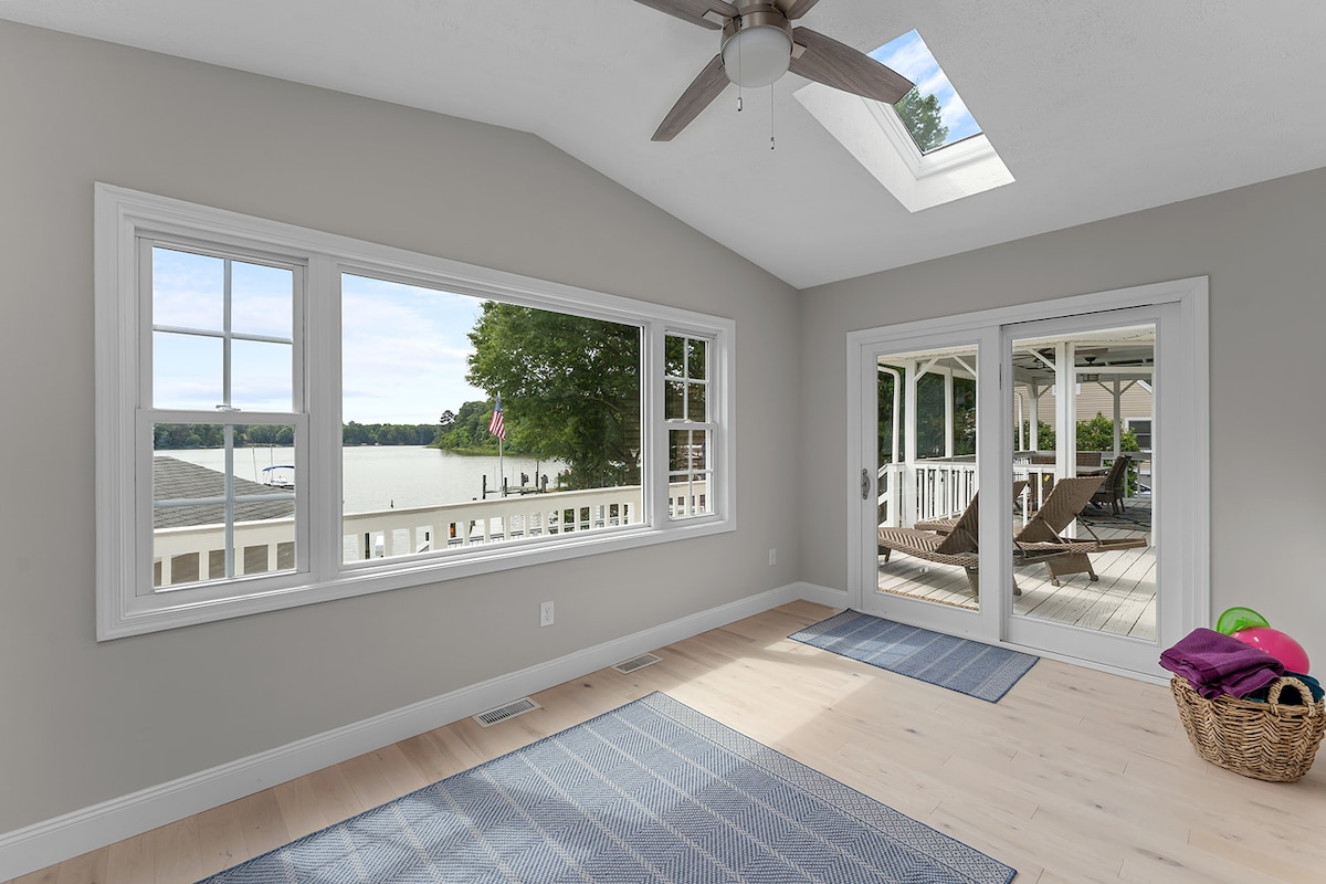 A bright room features large windows showcasing waterfront views. Natural light filters in through a skylight above, creating an airy atmosphere. Sliding doors provide access to the outdoor deck, with a smooth wooden floor and a soft area rug enhancing the space.
