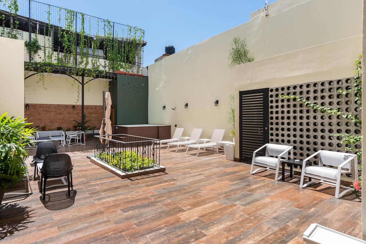An outdoor terrace is presented with a combination of lounge chairs and seating areas. Lush greenery and vertical plants add natural accents, while the textured flooring creates a warm surface. Sunlight illuminates the space, enhancing its open and inviting atmosphere.