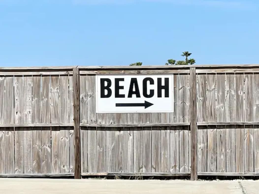 A wooden fence with a clear, large sign reading 'BEACH' directs towards the right. The sky above is bright and blue, creating a contrasting backdrop for the sign. The ground is paved, indicating a well-maintained pathway.