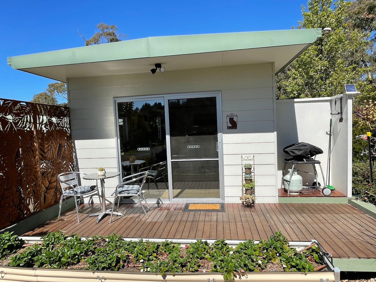 A modern retreat is presented with a deck area featuring two small chairs and a table. A BBQ grill is positioned nearby. Sliding glass doors lead into the interior, allowing natural light to brighten the space. Lush greenery and a garden border the deck.