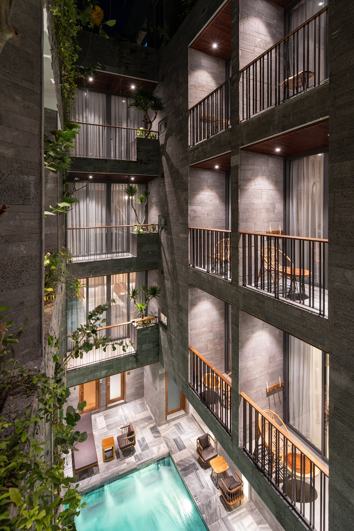 The villa's interior features multiple levels surrounding a serene pool area. Natural light filters through the glass doors and airy curtains, while greenery enhances the ambiance. Balconies provide seating with views of the pool, creating a relaxing environment.