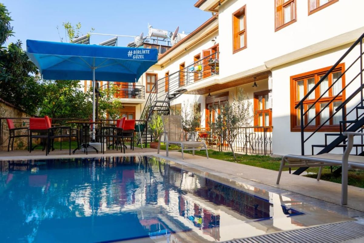 The image showcases a clear pool surrounded by a garden area. Bright patio furniture with a large umbrella is arranged on a wooden deck. The two-story building features multiple windows with wooden frames, and stairs leading to the upper level are visible.