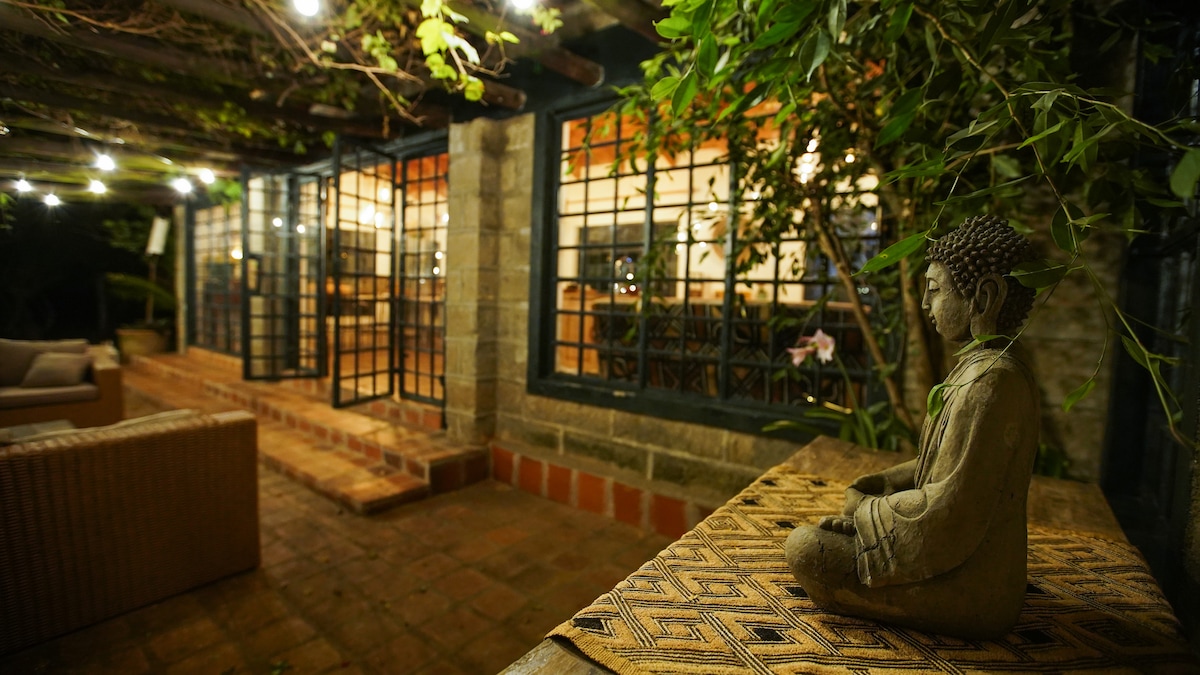 A serene outdoor seating area is framed by lush greenery and soft ambient lighting. A stone Buddha statue rests on a patterned table, offering a peaceful focal point. The glass doors of the cottage are partially visible, leading to an inviting interior space.