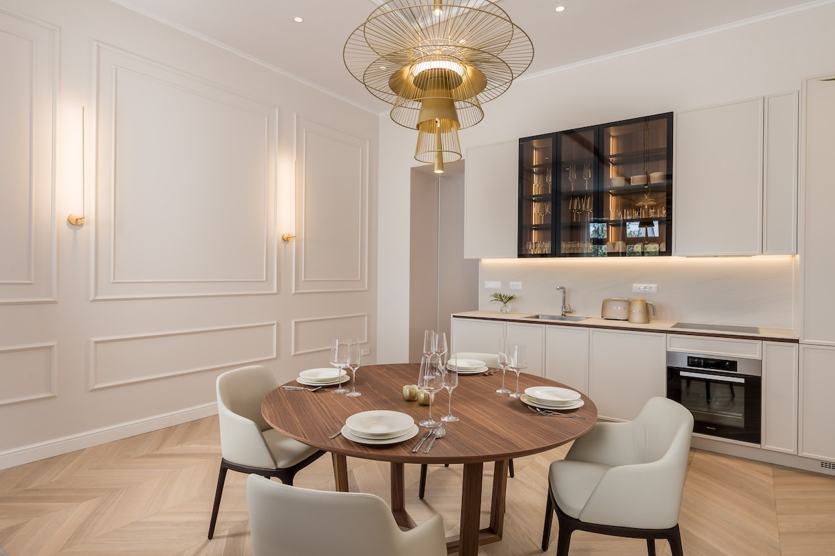 A dining area features a round wooden table set for four, with simple white plates and tall glassware. Stylish white dining chairs surround the table. An elegant light fixture hangs from the ceiling, and a sleek kitchen area with cabinetry and appliances is visible in the background.