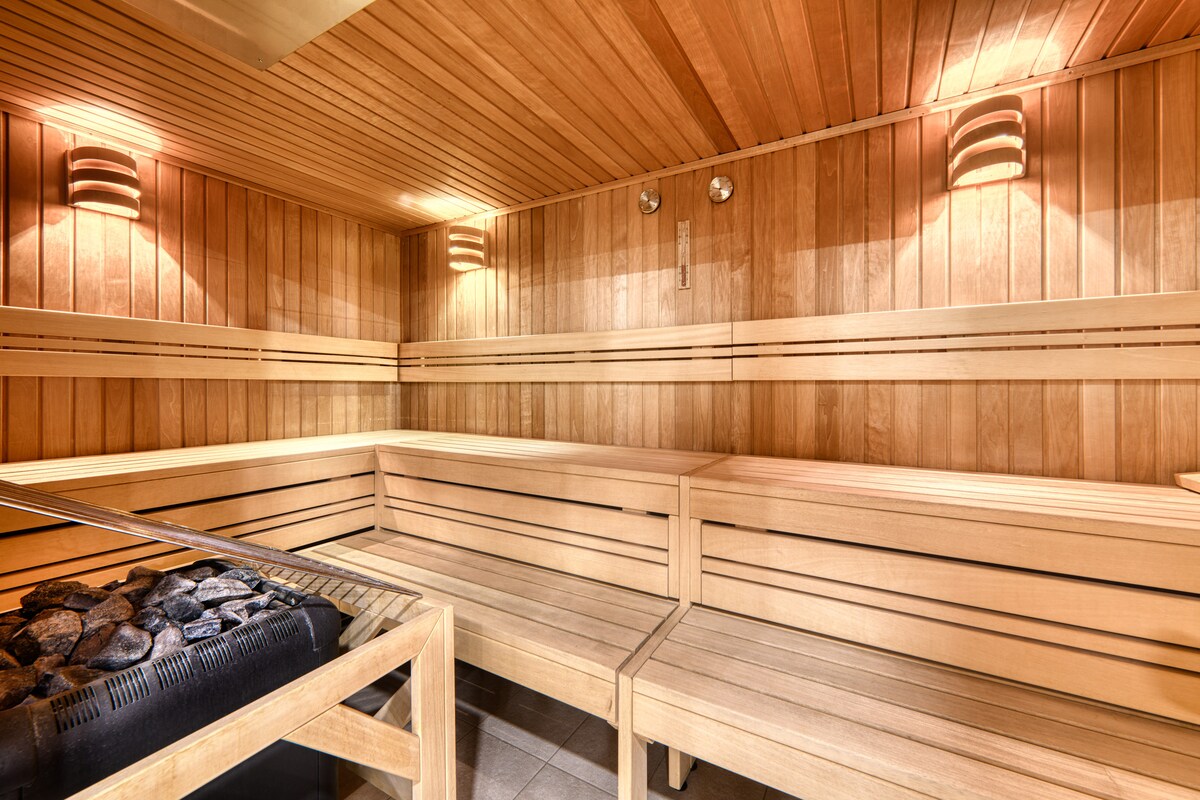 The sauna area features warm wooden benches arranged in a spacious layout, designed for relaxation. A stone heater is positioned at one end, contributing to the tranquil atmosphere created by soft, indirect lighting along the walls.
