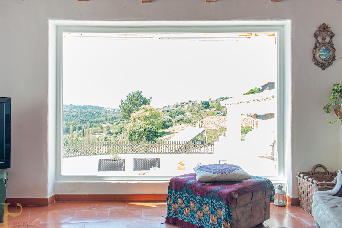 A large window frames a view of the surrounding greenery and hills, allowing natural light to fill the room. A colorful blanket drapes over a spacious ottoman in front of the window, enhancing the inviting ambiance. The peaceful outdoor scenery is clearly visible.