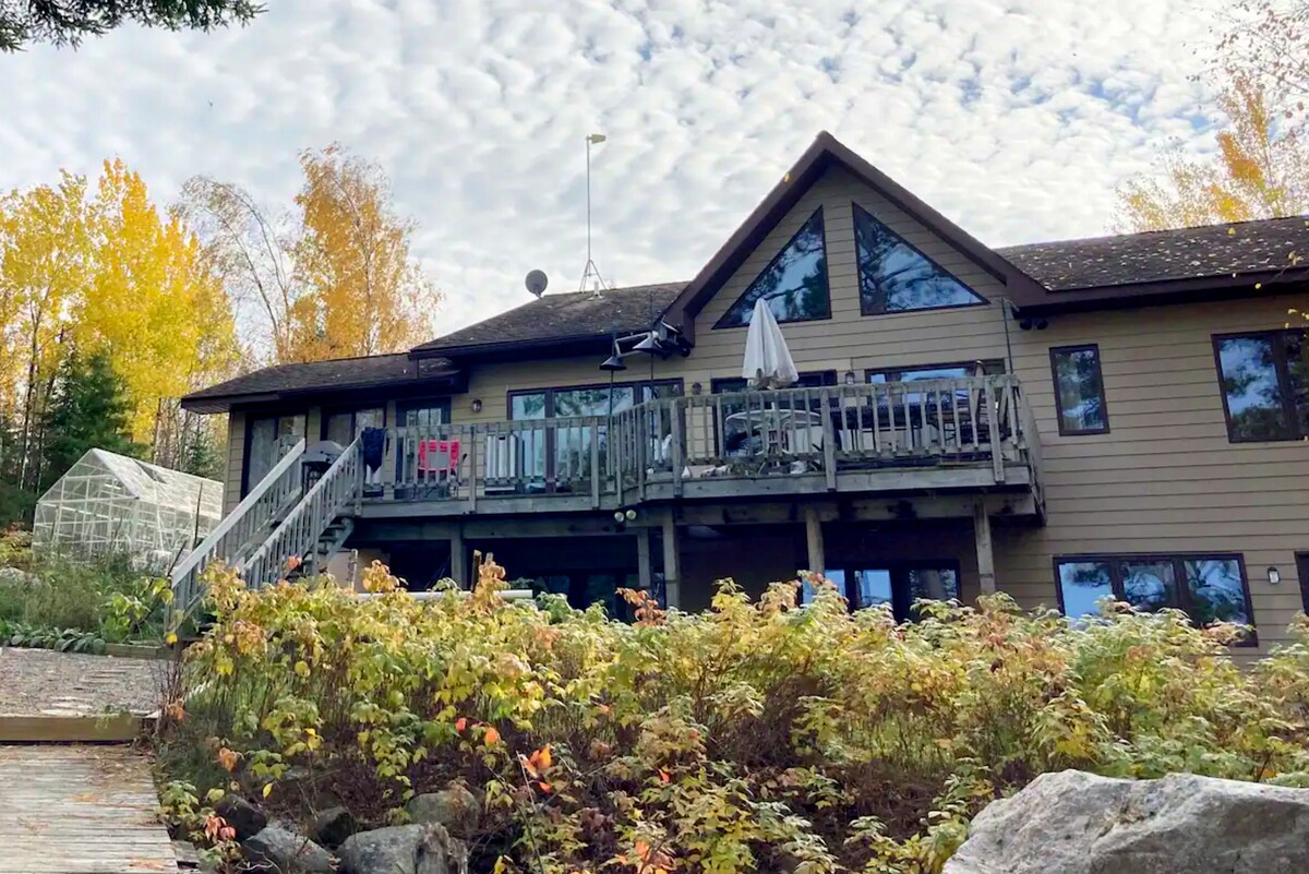 The cabin exterior features a sloped roof and large windows that provide a view of the surrounding landscape. A spacious deck is visible, adorned with a patio umbrella and colorful chairs. Flanking the approach are blooming plants and shrubs, reflecting autumn colors.
