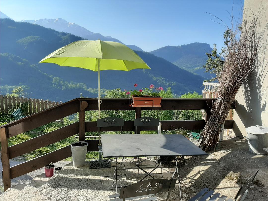 A terrace area is presented with a light green umbrella providing shade over a metal table and chairs. A planter with flowers and scenic mountain views create a peaceful outdoor space. Natural elements are visible in the surrounding landscape.