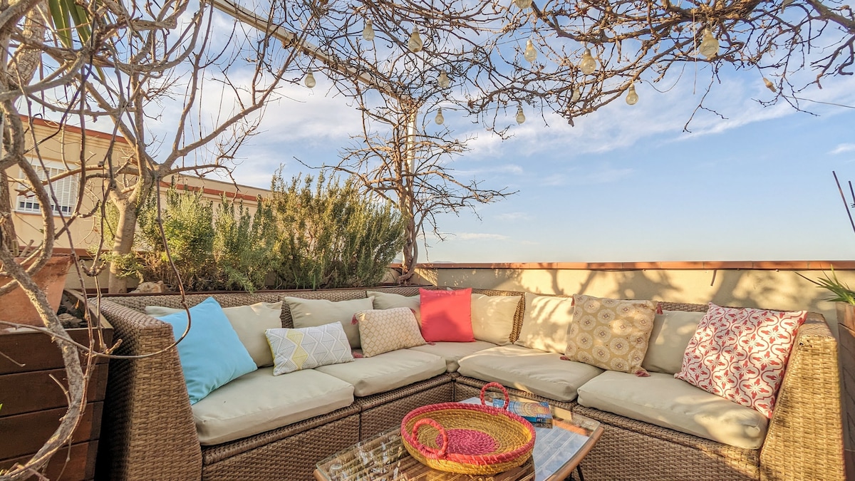 A spacious terrace is adorned with a vine-covered pergola, providing shade and a relaxing atmosphere. A large, circular seating area is arranged with neutral cushions in various colors. A woven basket and several books are placed on a glass-top table, inviting leisurely moments.