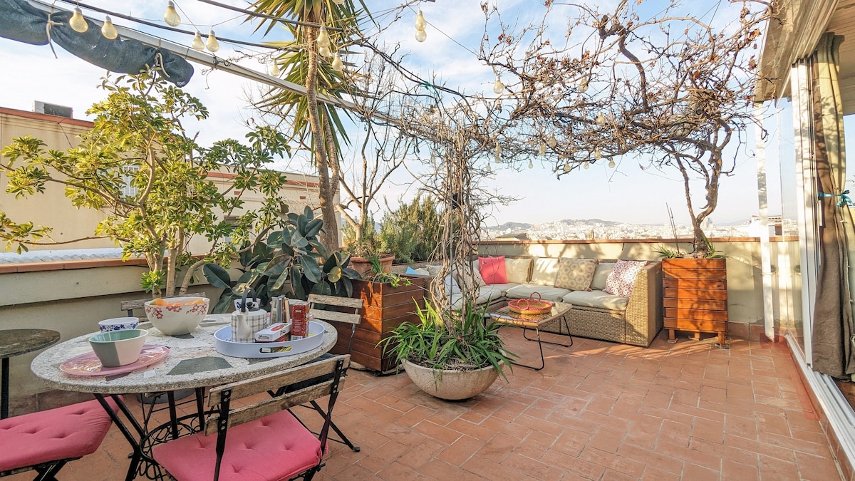 A spacious terrace features a round table surrounded by pink chairs, with a lounging area furnished with cushions. Green plants and a grape arbor add natural charm, while string lights create a cozy atmosphere. The view extends across the city skyline.