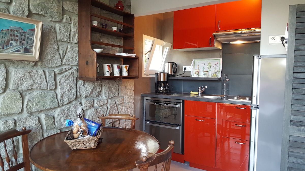 A compact kitchen area features bright orange cabinetry paired with dark countertops. Wooden shelving holds dishware and decorative items. A round table with chairs occupies the space, while a window allows natural light to brighten the room.