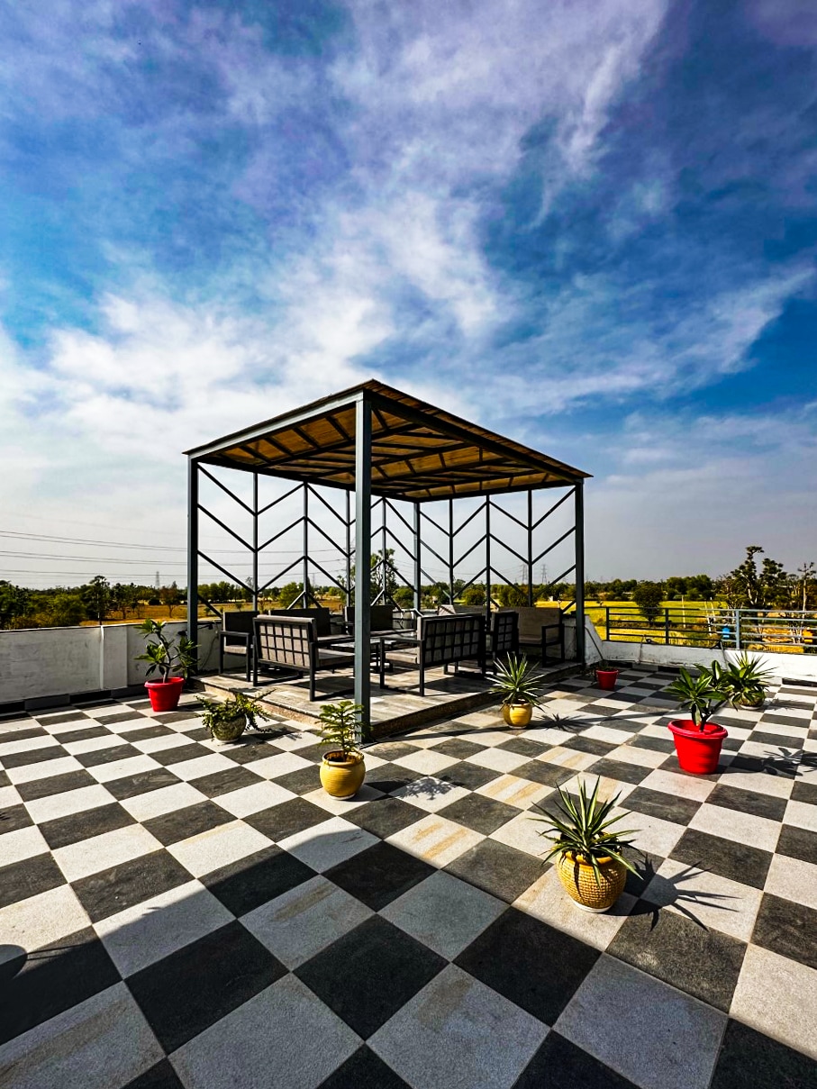 A spacious rooftop area features a canopy structure supported by a sleek frame. Comfortable seating is arranged beneath the cover, surrounded by potted plants in vibrant red and neutral tones. A checkerboard pattern adorns the floor, enhancing the open sky backdrop.