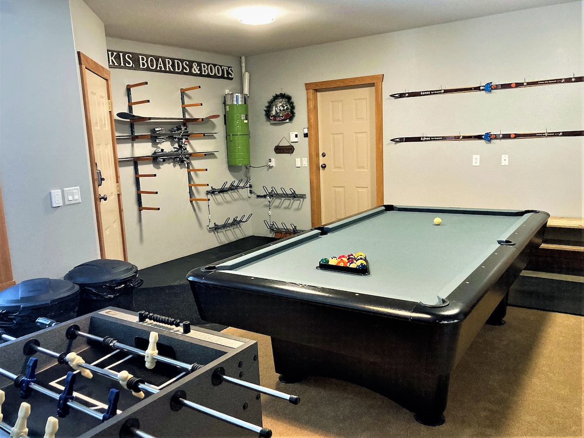 A dedicated games room is showcased with a pool table and foosball table for entertainment. Ski and snowboard equipment is organized on wall racks, with ample storage available. The space includes a cozy carpeted area, enhancing the functionality for leisure activities after a day on the slopes.