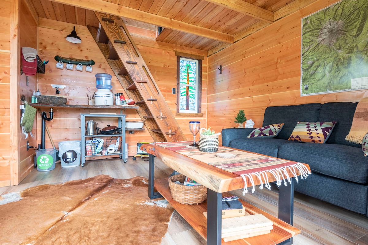 The inviting interior showcases warm wooden walls and ceiling. A cozy seating area features a dark love seat, a rustic coffee table, and a decorative animal hide on the floor. A compact kitchenette area is equipped with a rock sink and practical storage furniture, with a staircase leading to a loft.