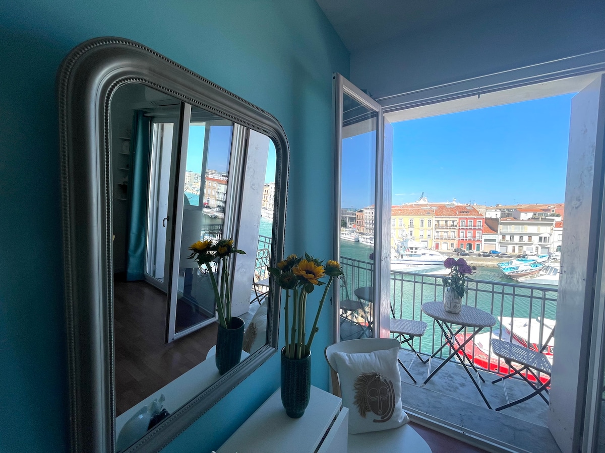 A room featuring a large mirror reflects fresh sunflowers in a vase. Double French doors open to reveal a vibrant view of colorful boats along the turquoise water, framed by a cheerful blue wall and a light-colored cushioned chair beside a small table.
