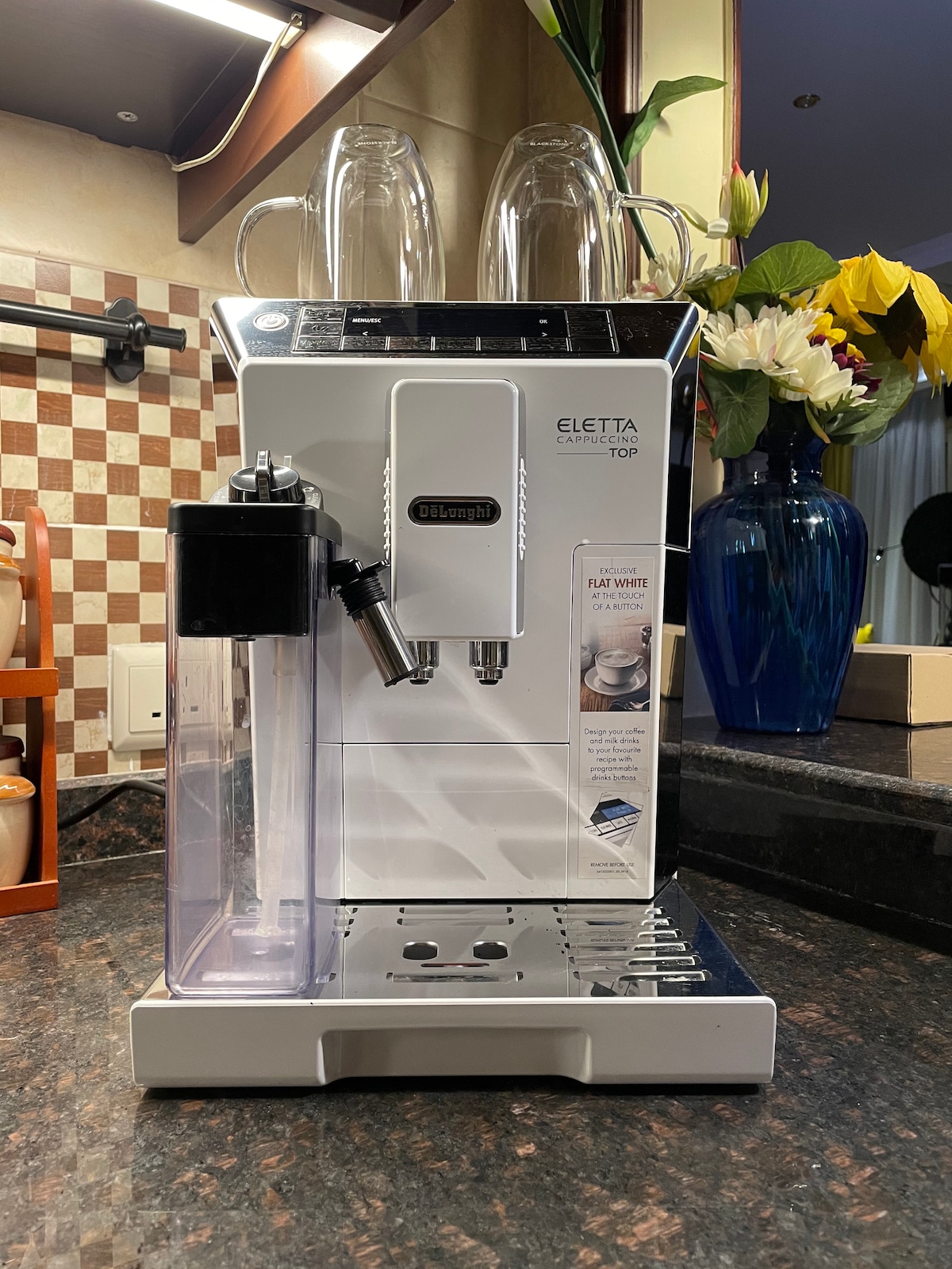 A fully automated coffee machine is positioned on a granite countertop. It features a sleek design with a brewing unit, a water reservoir on the side, and space for two cups above. Surrounding elements include a checkered backsplash and a flower vase, adding to the kitchen aesthetic.